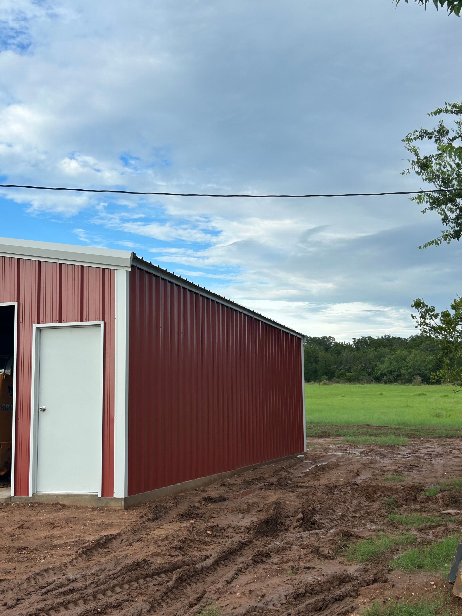 Red metal shed with white door beside muddy ground and green field under a cloudy sky