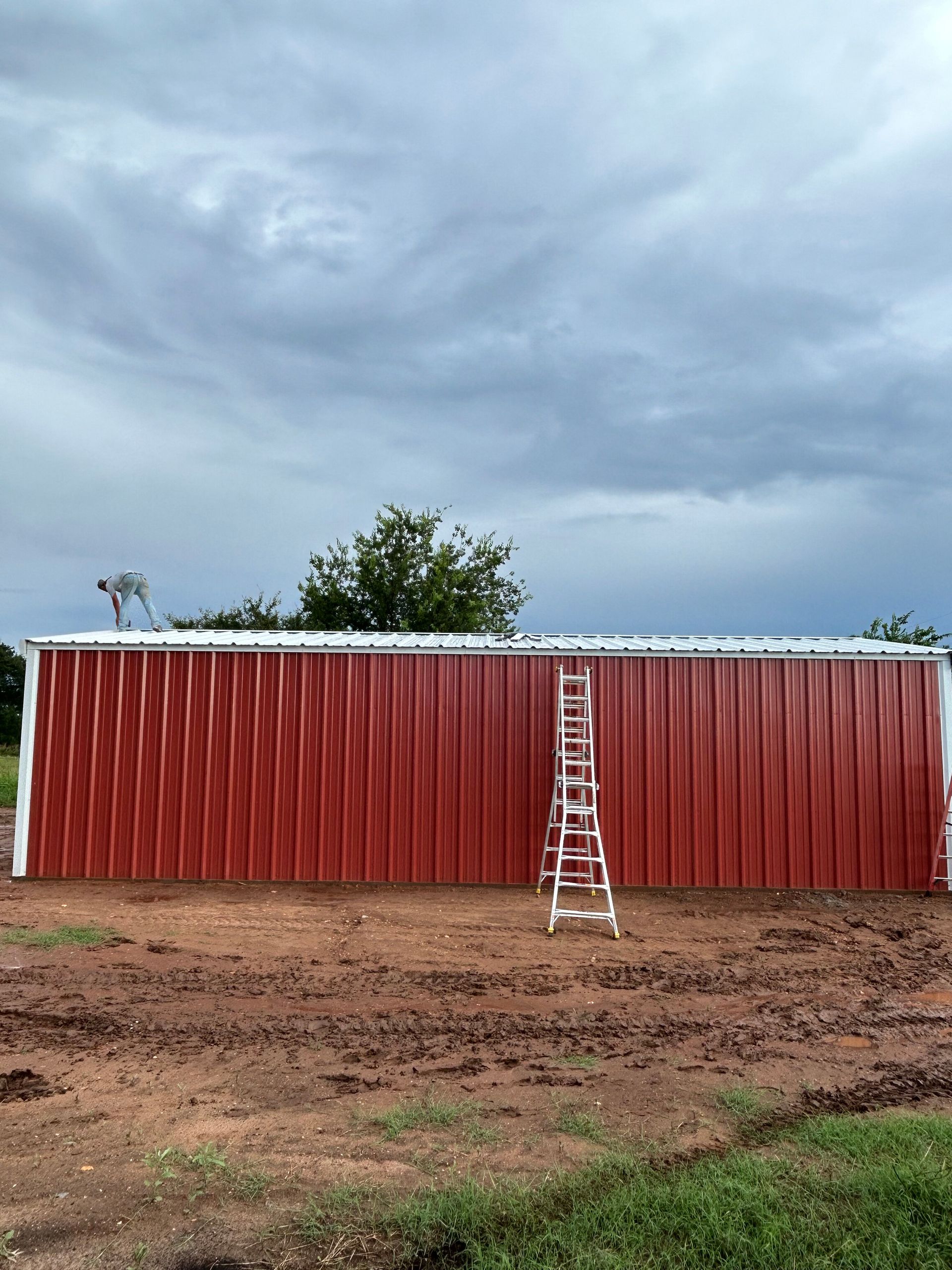 Red barn with a ladder leaning against it under a cloudy sky on a dirt lot.