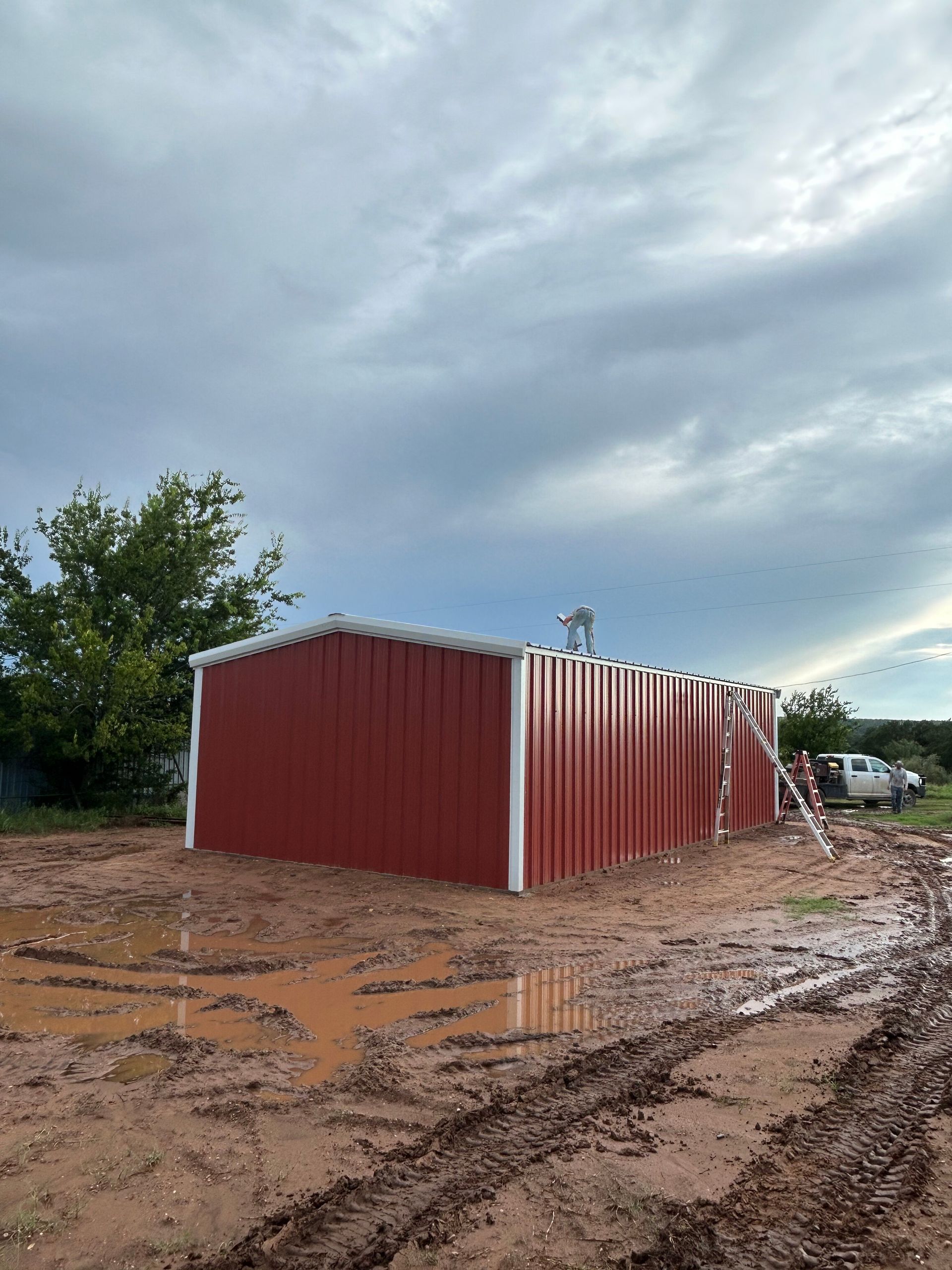 Red shipping container on muddy ground under a cloudy sky, with trees and a vehicle in the background.