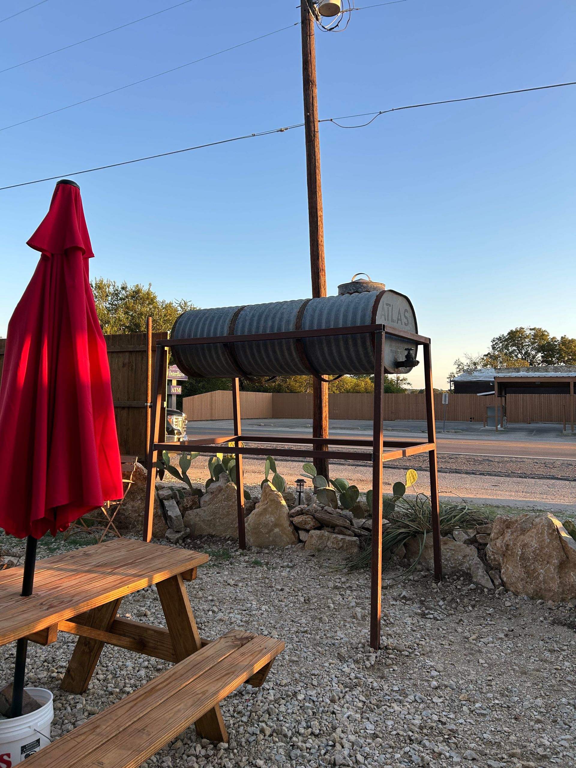Red patio umbrella and picnic table beside a gravel yard with a metal grill under a clear blue sky.