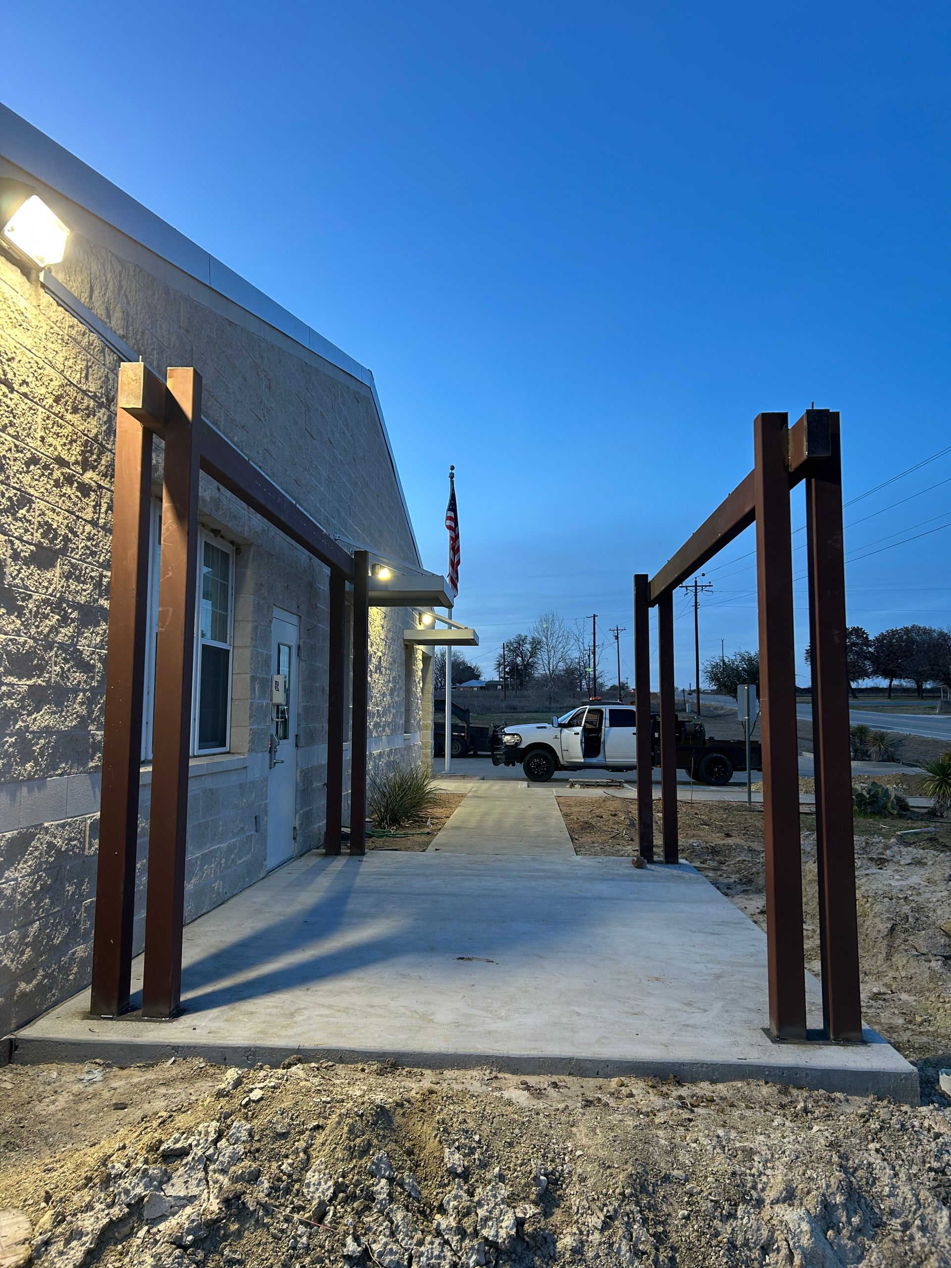 Covered concrete patio beside a stone building at dusk, with rusty metal posts and a parked vehicle in the background