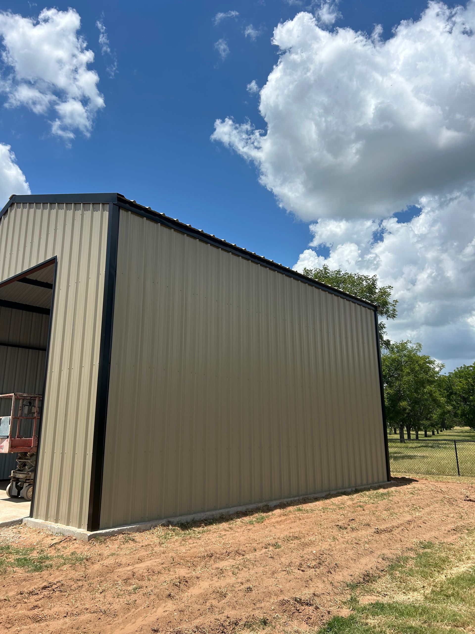 Beige metal shed on a dirt lot under a blue sky with clouds, near trees and open fields
