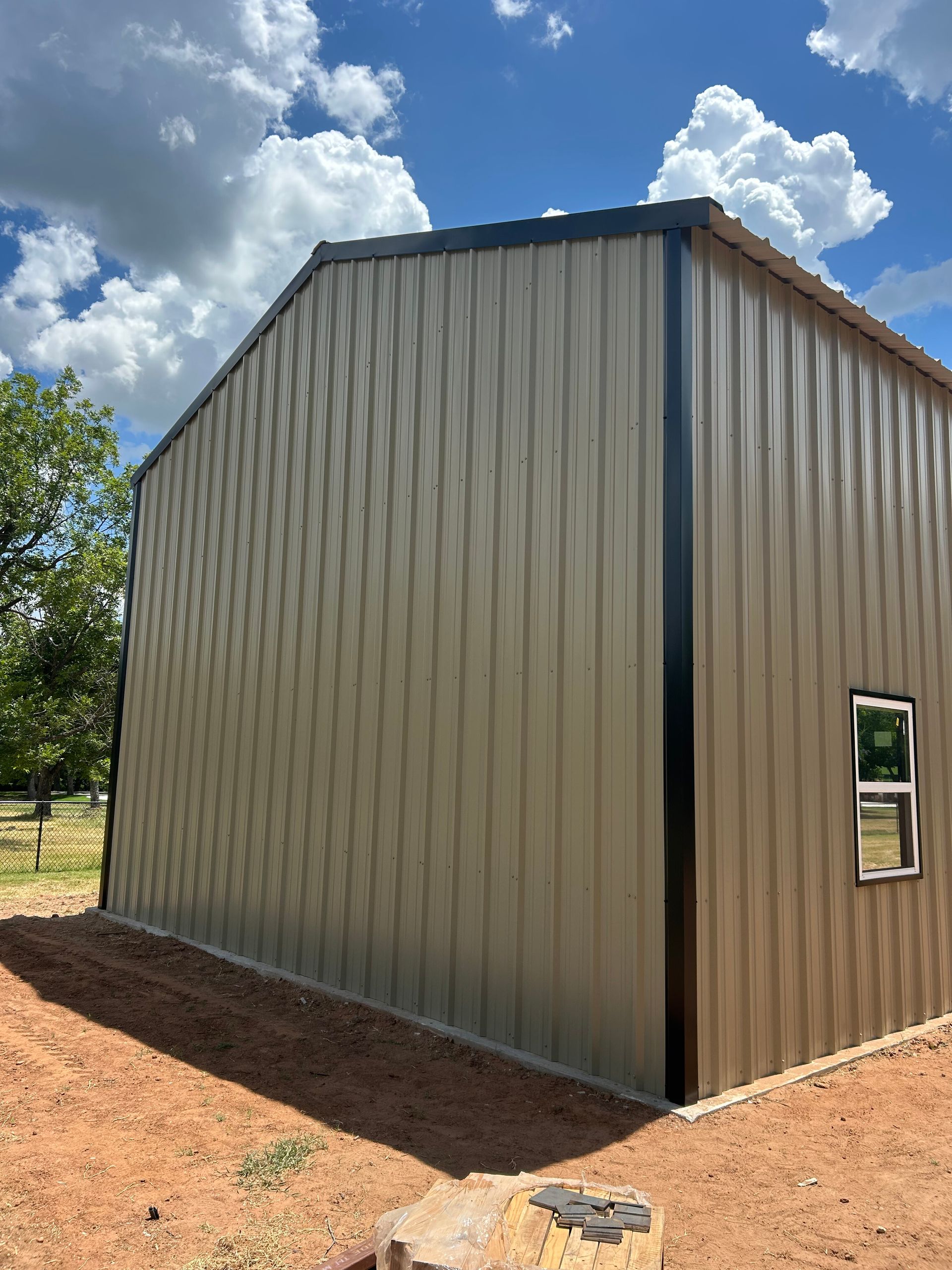 Small tan metal shed with blue roof trim and a window, standing on red dirt under a cloudy sky.