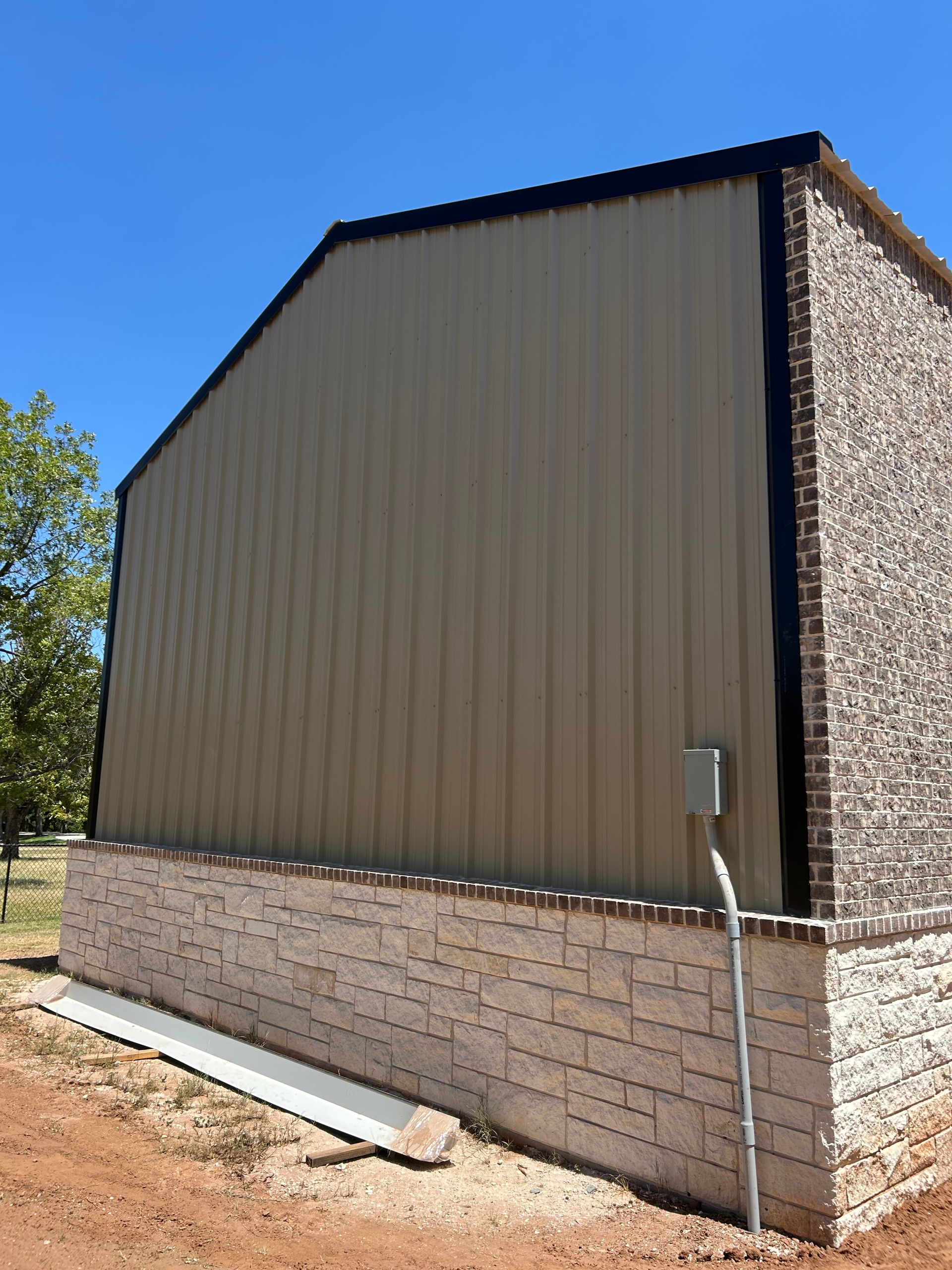 Tan metal-sided outbuilding with brick base and electrical conduit, seen from the side under a blue sky