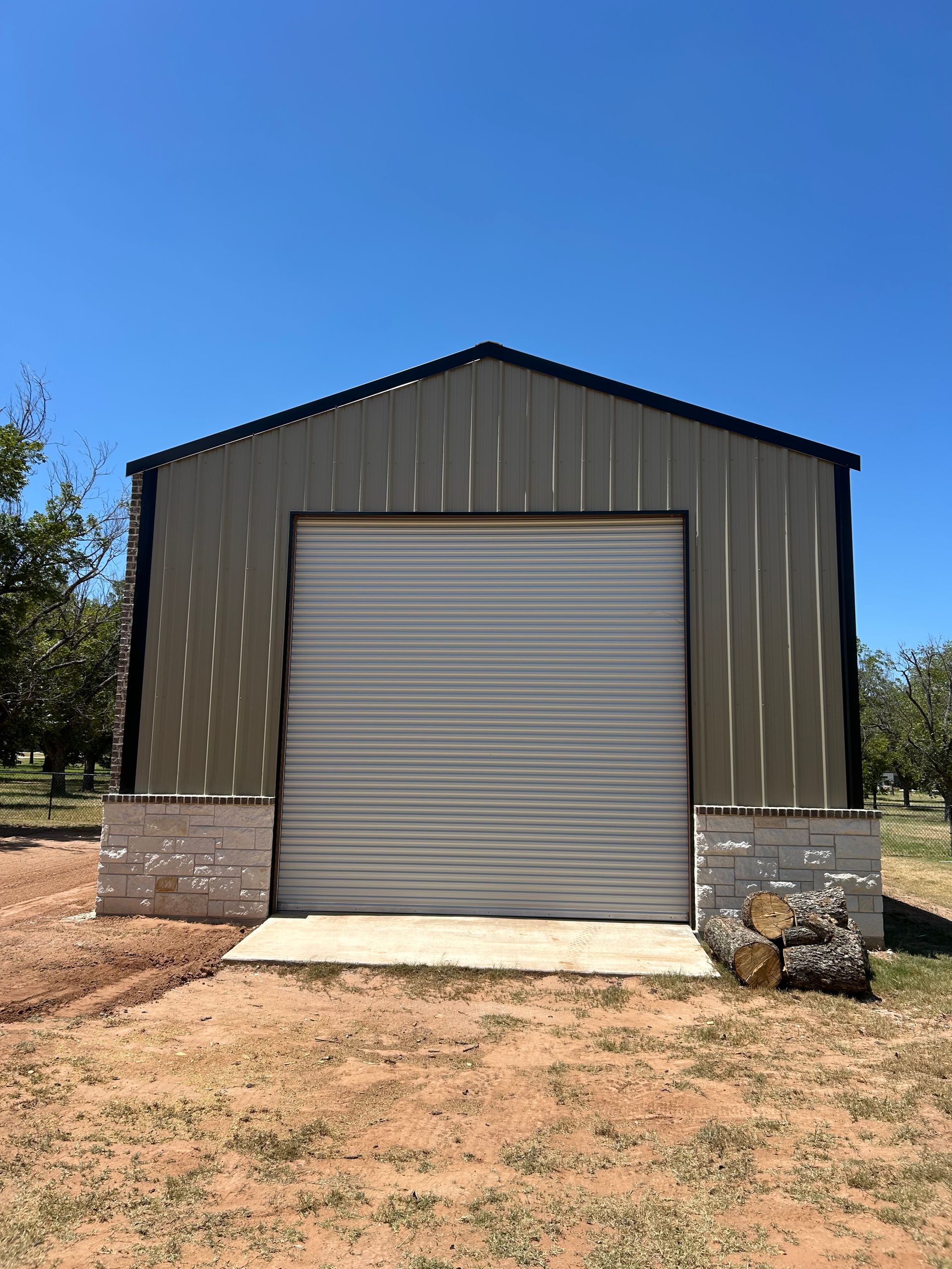 Metal shed with a closed gray roll-up door, tan stone base, and dirt yard under a blue sky