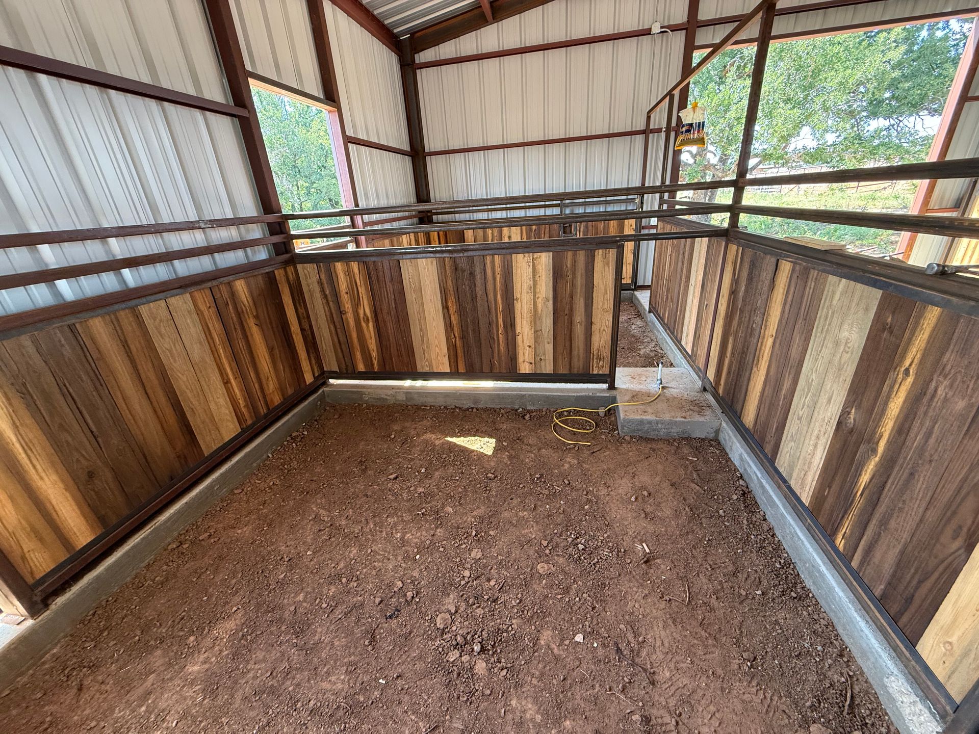 Small empty livestock pen with dirt floor, wooden walls, and a corrugated metal roof.