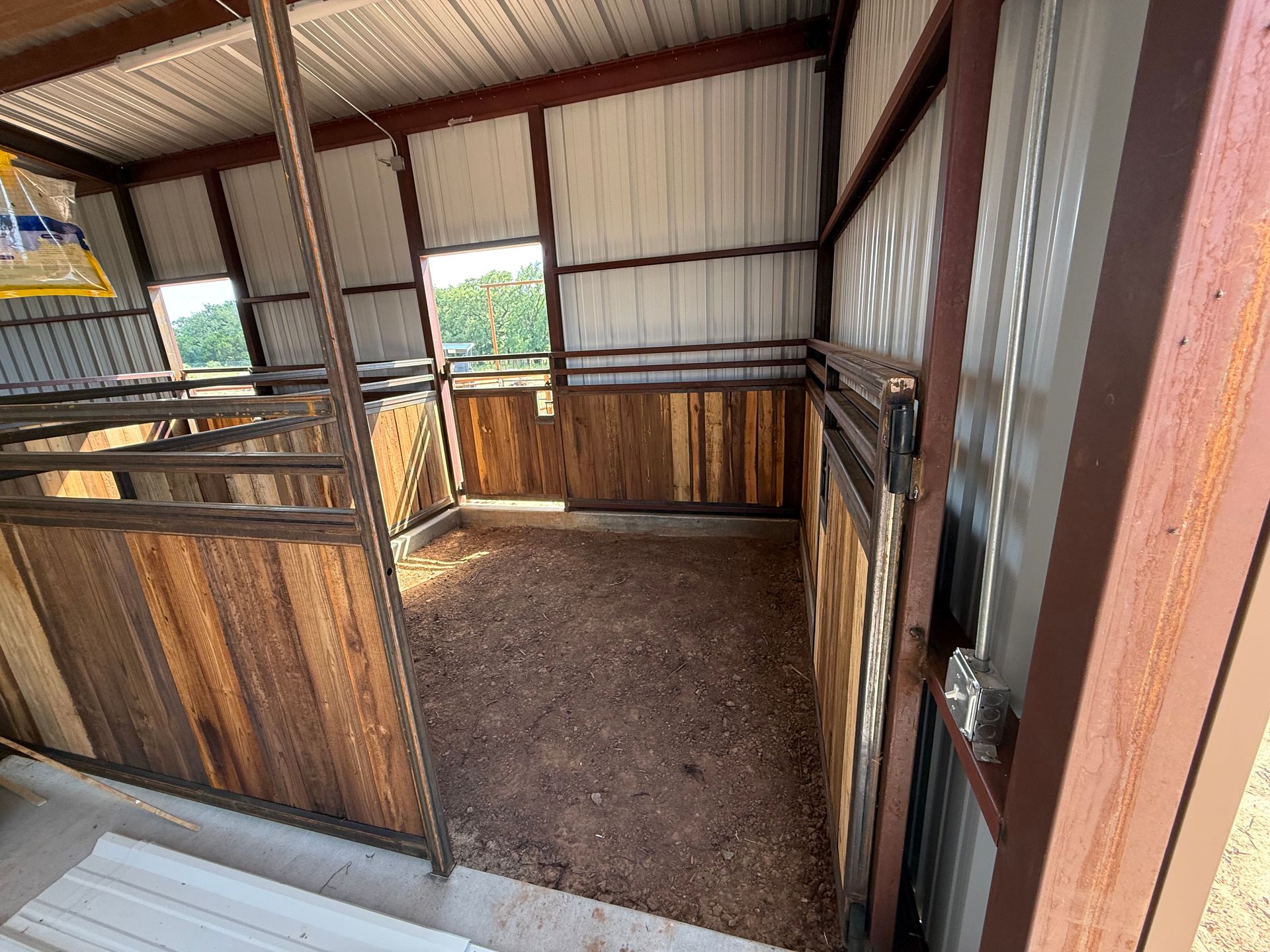 Empty horse stall with wood-paneled walls, metal rails, and dirt floor inside a barn.