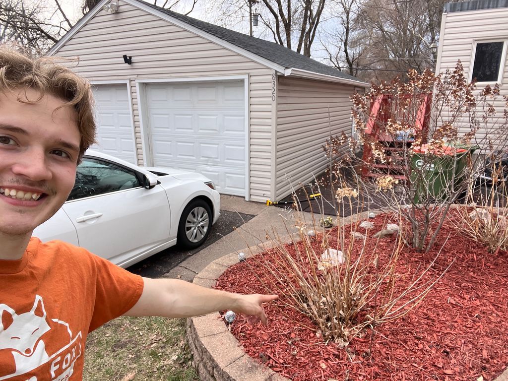 Smiling man in orange shirt pointing at red mulch garden beside a driveway and garage, with white car nearby