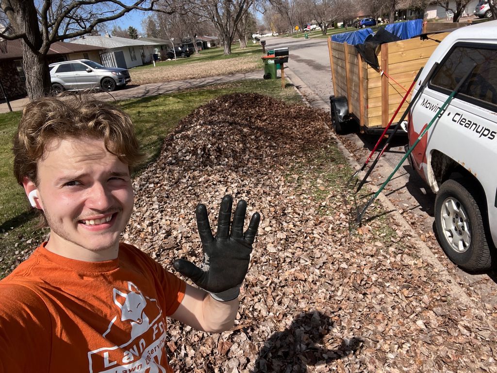 Person in orange shirt waving in a leaf-covered yard beside a truck and wood fence