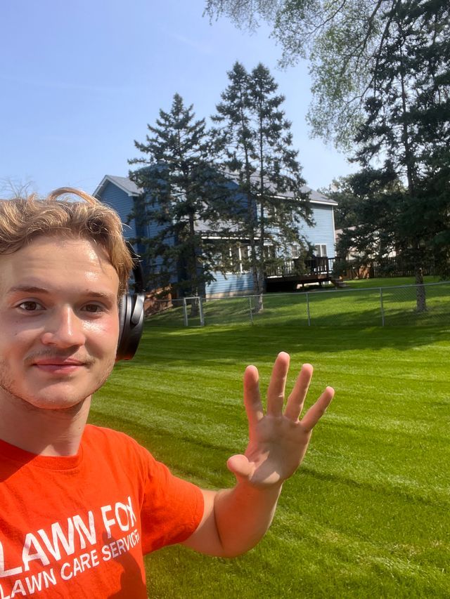 Selfie of a person in an orange shirt waving on a sunny lawn with trees and a building behind.