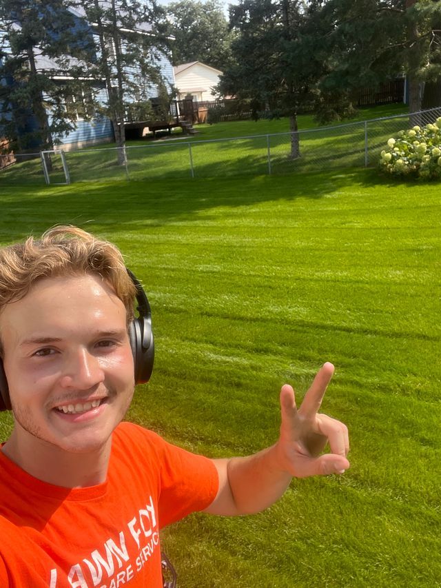 Person in orange shirt taking a selfie in a sunny green yard, smiling and making a hand sign.