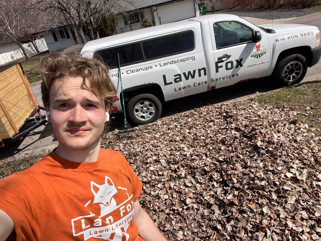 Selfie of a person in an orange shirt in front of a white Lawn Fox service truck and autumn leaves.