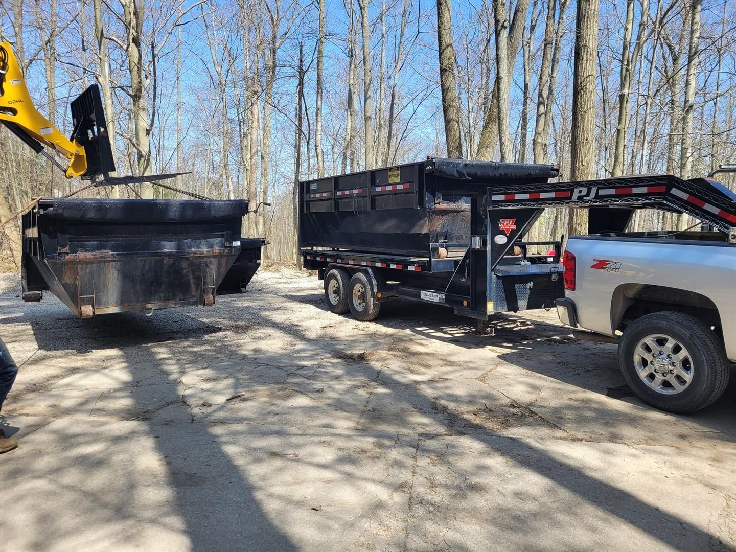 A skid steer loading a dumpster onto a trailer hitched to a truck outdoors on a sunny day.