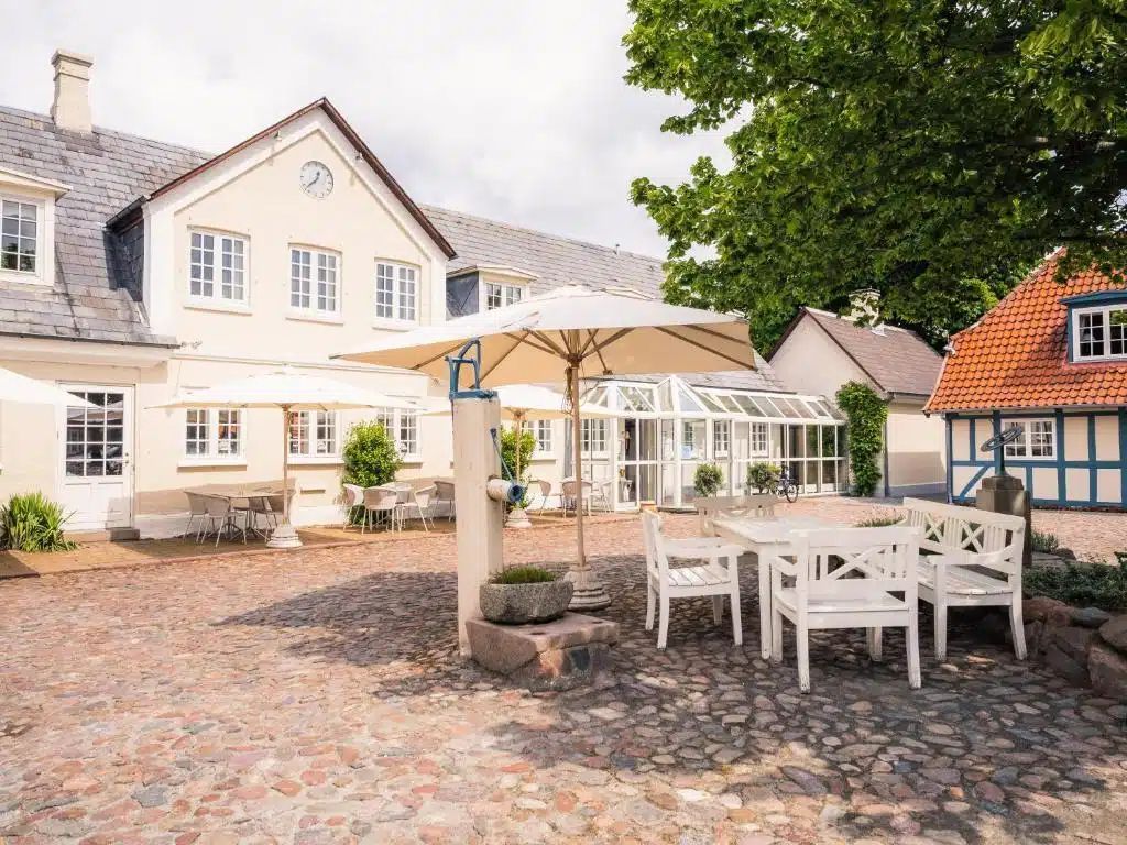 Ydersiden af ​​en bygning med udendørs siddepladser og en glasindkapslet struktur på en brostensbelagt terrasse.