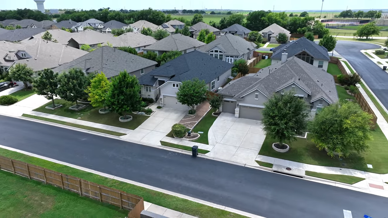 Aerial view of a suburban neighborhood with rows of houses, driveways, trees, and a road in front.