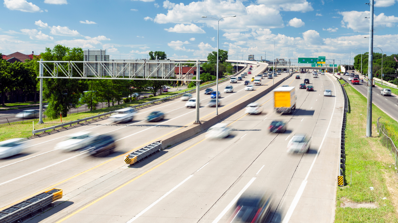 Busy multi-lane highway with cars and trucks under a bright blue sky, viewed from an overpass