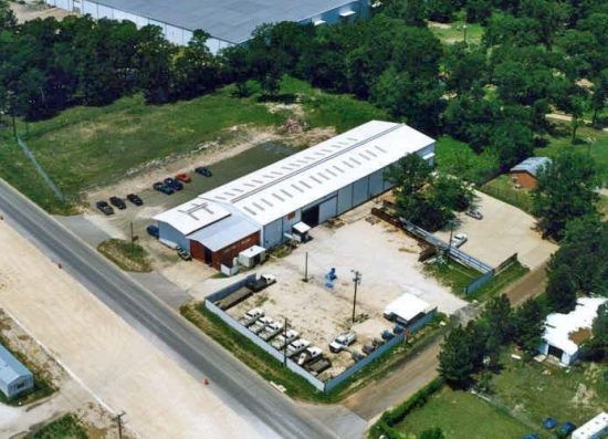 Aerial view of an industrial building with a long white roof, surrounded by trees and a paved area.