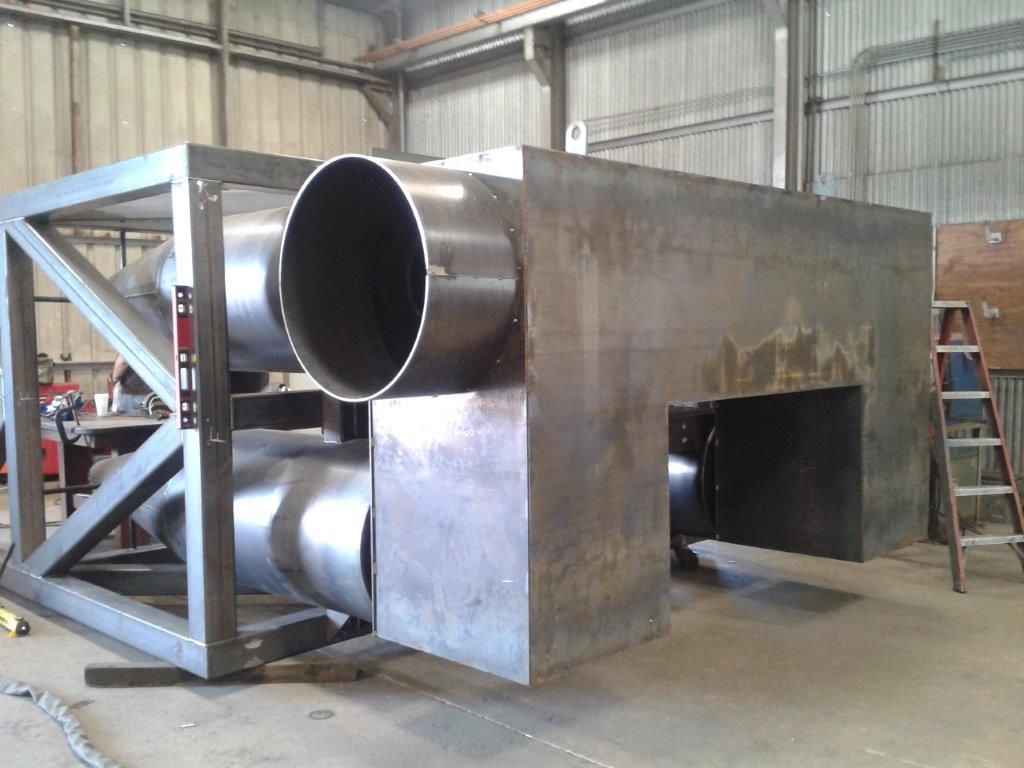 Large metal industrial component with cylindrical ducts, supported by a steel frame, inside a workshop.