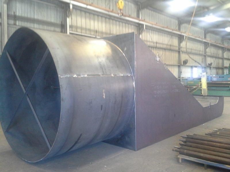 Large metal ductwork component inside a factory; cylindrical section with internal fins attached to a rectangular section.