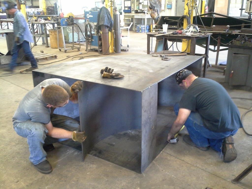 Three people working on a large, metal box-shaped structure in a workshop.