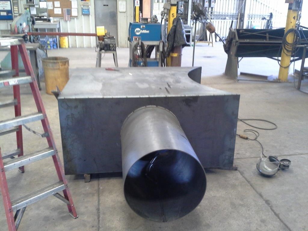 Large metal box with cylindrical pipe attached in a workshop setting; a welding machine is in the background.