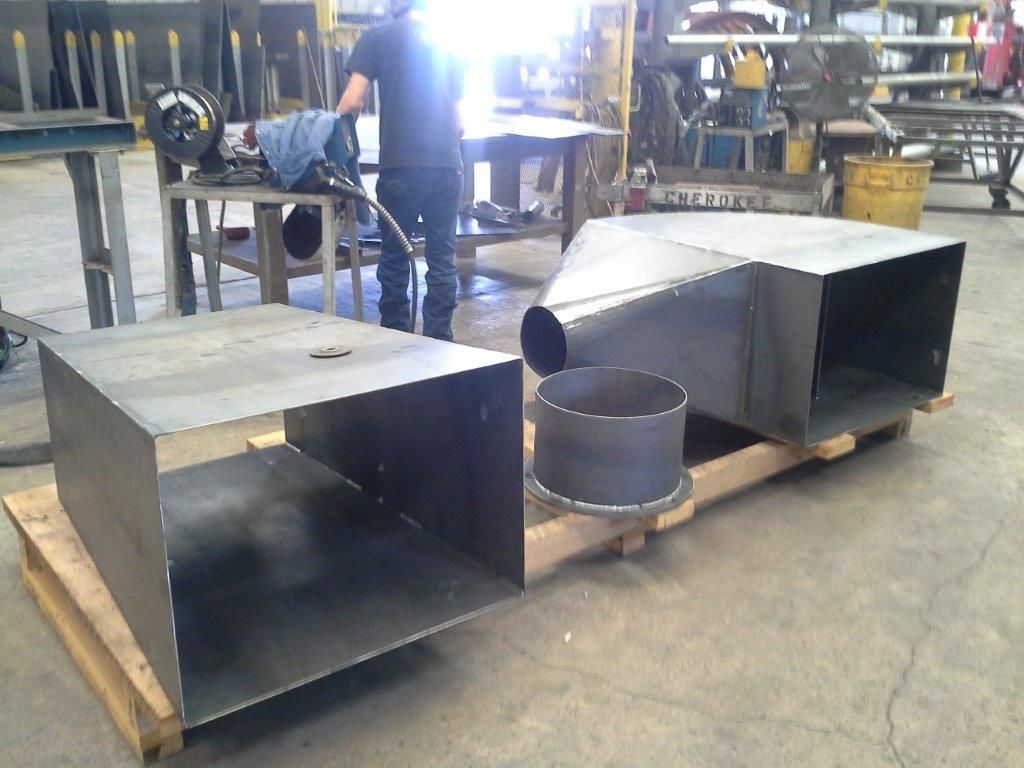 Metal fabrication shop: Fabricated rectangular ductwork, cylindrical pipe, and a worker welding in the background.
