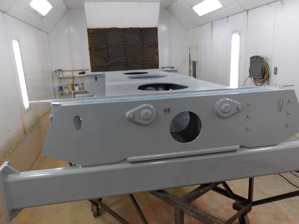 A large, gray metal object, possibly a tank hull, being painted in a spray booth.