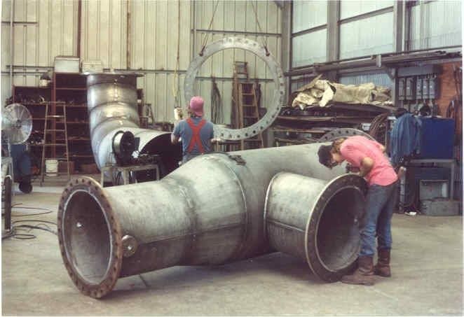 Workers inspecting a large industrial pipe assembly in a workshop; a flange hangs in the background.