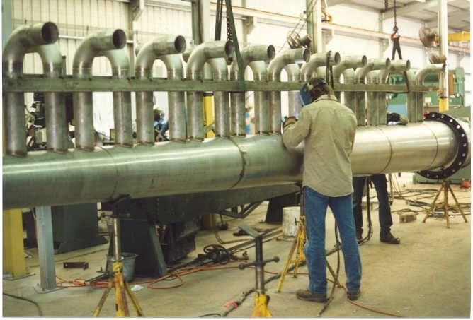 A worker welds pipes in a factory setting. Large stainless steel pipe with multiple smaller pipes attached.