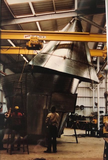 Large metal structure being assembled in a workshop, crane overhead, workers observing.