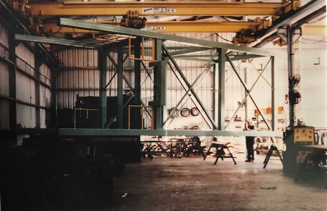 Large metal frame suspended from overhead crane in a workshop; person standing nearby.