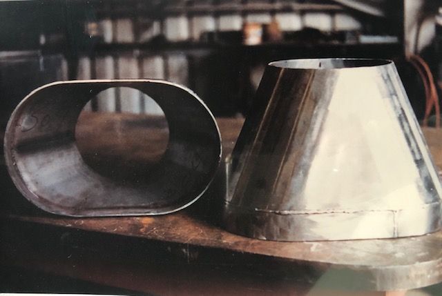 Two metal exhaust components: an oval-shaped pipe and a cone-shaped section. They sit on a metal surface.
