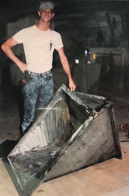 Man in workshop holding large, trapezoidal metal container; industrial setting.
