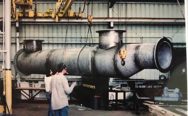 Large metal industrial pipe suspended by cranes, two men looking up at it in a factory setting.