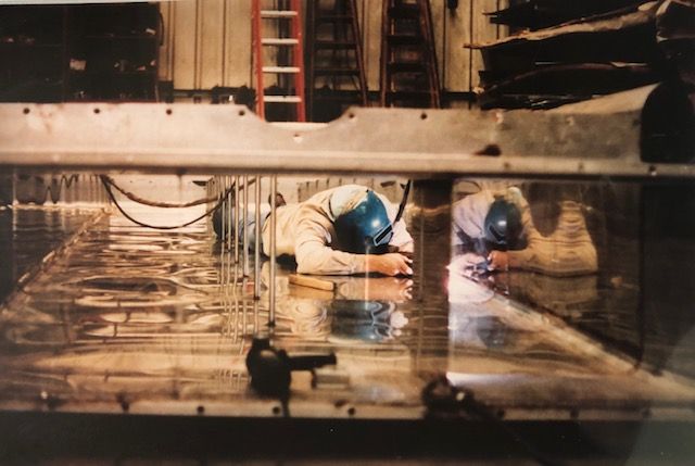 Two workers welding inside a metal tank, with their reflections visible, wearing helmets.