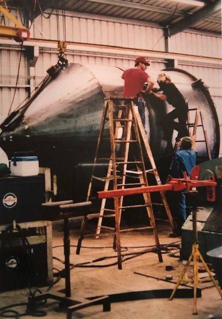 Workers on ladders, assembling large metal tank inside a workshop.