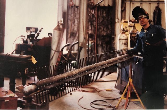 Man welding a large metal structure in a workshop. He wears a welding mask and gloves, working near equipment and tanks.