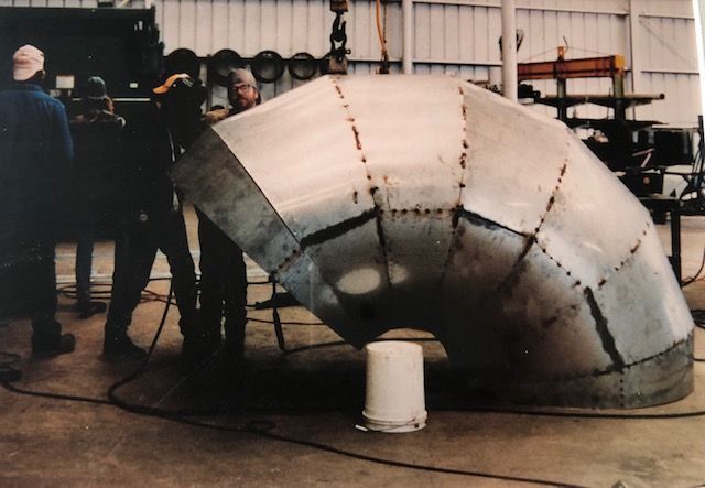 Large metal elbow pipe being constructed inside a factory, several workers present.