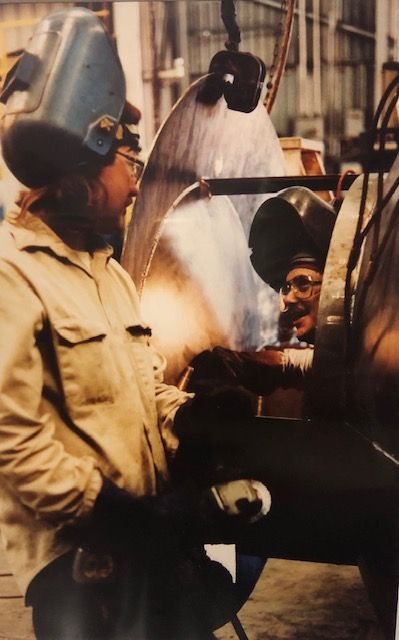 Two welders with helmets working on large metal sculpture. One looks at the other. Indoor setting.