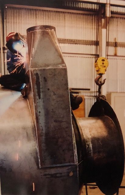 Welder works on large metal structure inside a warehouse, with sparks and a crane hook visible.