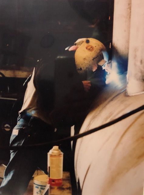 A person welds metal in a dim setting, with bright sparks and a protective mask.