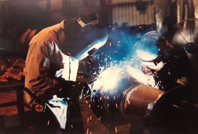 Welder in a workshop, sparks flying, welding metal pipe, another welder in the background.