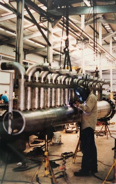 Welder working on large industrial pipe structure in a factory; overhead crane in use.