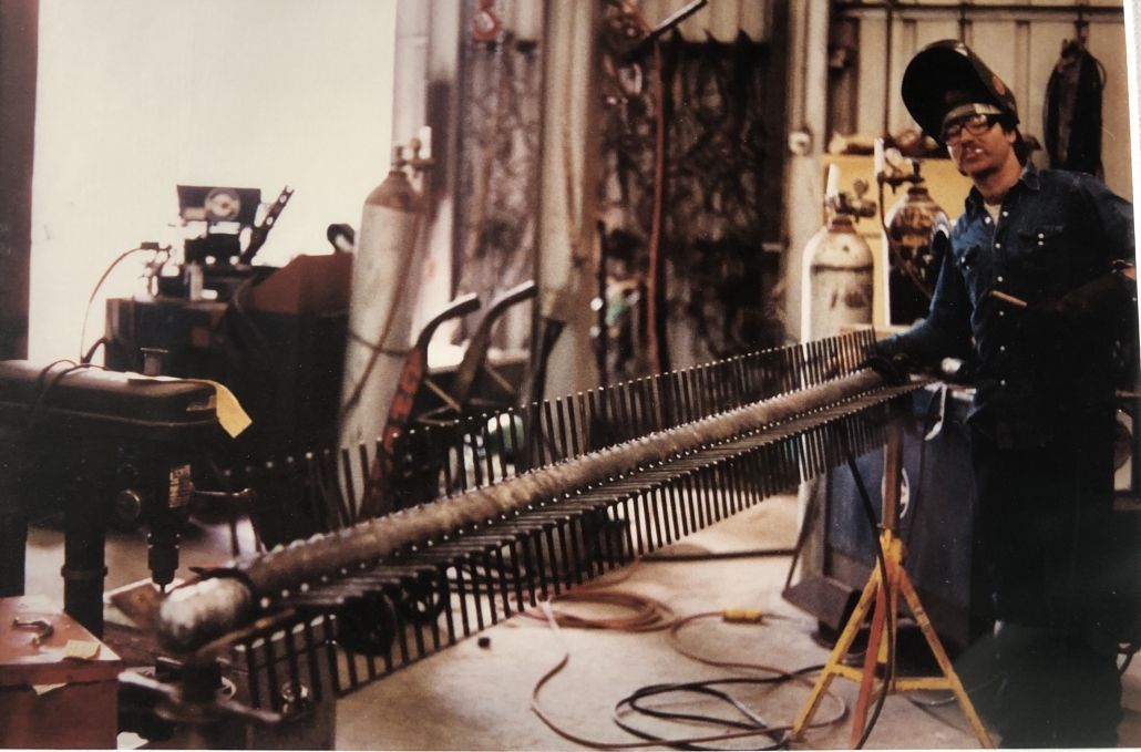 Welder in workshop, wearing protective gear, working on a large metal structure with numerous rods.