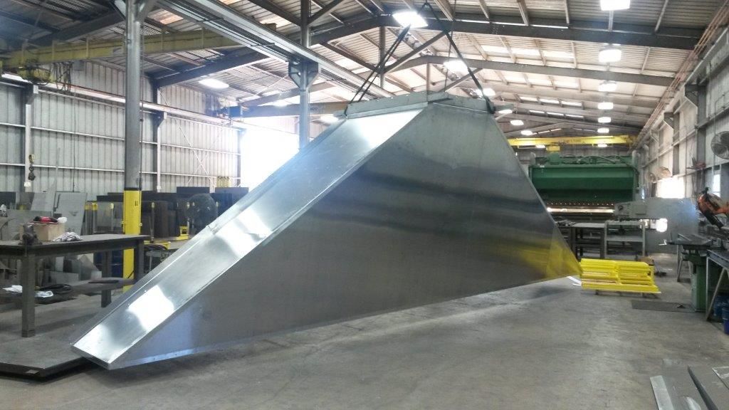 Large, angled, metallic structure suspended by a crane in an industrial warehouse.