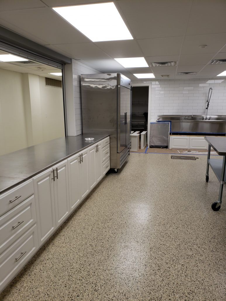 Commercial kitchen with stainless steel appliances and cabinets, light-colored speckled flooring, and a long counter.