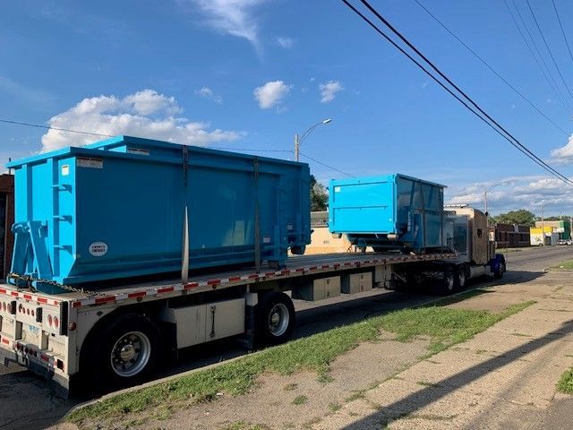 A semi-truck parked on a street carries two bright blue industrial dumpsters on its flatbed trailer.