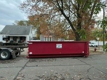 A large, red dumpster sits on a residential street in front of a house, attached to a truck trailer.