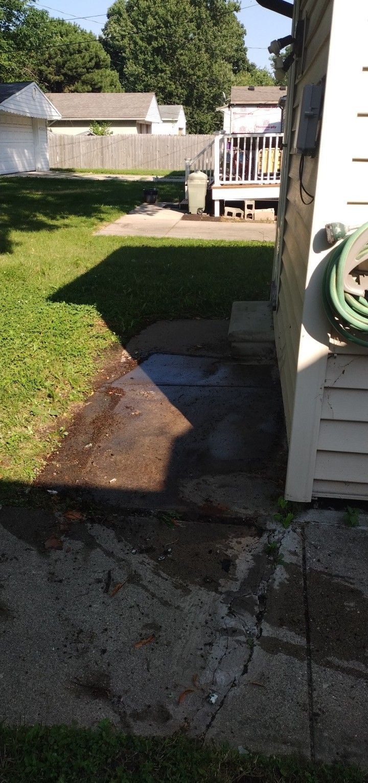 A side yard view showing a concrete pathway leading toward a wooden deck and a green trash bin in a grassy backyard.