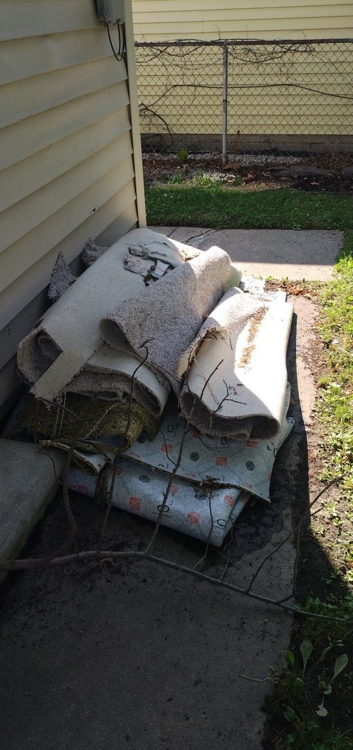 A pile of rolled-up, weathered carpet and padding rests on a concrete walkway beside the side of a house.